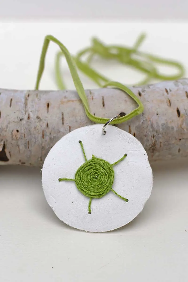 woven web embroidered clay pendant in green resting on log with green cord